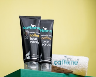 Two black tubes of mCaffeine naked and raw coffee face wash and face scrub are placed on a green surface against a yellow background. Beside them is a white towel with 'mCaffeine' embroidered in blue and a small wooden spatula.