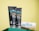 Two black tubes of mCaffeine naked and raw coffee face wash and face scrub are placed on a green surface against a yellow background. Beside them is a white towel with 'mCaffeine' embroidered in blue and a small wooden spatula.