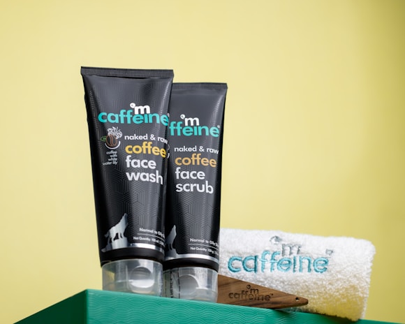 Two black tubes of mCaffeine naked and raw coffee face wash and face scrub are placed on a green surface against a yellow background. Beside them is a white towel with 'mCaffeine' embroidered in blue and a small wooden spatula.