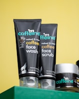 Close-up of three skincare products from the brand mCaffeine placed on a green surface against a yellow background. Two black tubes labeled 'naked & raw coffee face wash' and 'coffee face scrub' are prominently displayed along with a round container, also branded.