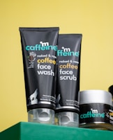 Close-up of three skincare products from the brand mCaffeine placed on a green surface against a yellow background. Two black tubes labeled 'naked & raw coffee face wash' and 'coffee face scrub' are prominently displayed along with a round container, also branded.