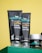 Close-up of three skincare products from the brand mCaffeine placed on a green surface against a yellow background. Two black tubes labeled 'naked & raw coffee face wash' and 'coffee face scrub' are prominently displayed along with a round container, also branded.