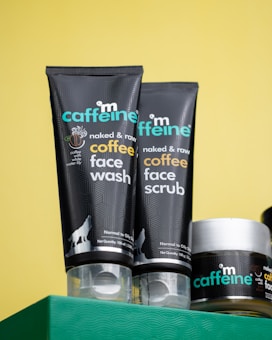 Close-up of three skincare products from the brand mCaffeine placed on a green surface against a yellow background. Two black tubes labeled 'naked & raw coffee face wash' and 'coffee face scrub' are prominently displayed along with a round container, also branded.