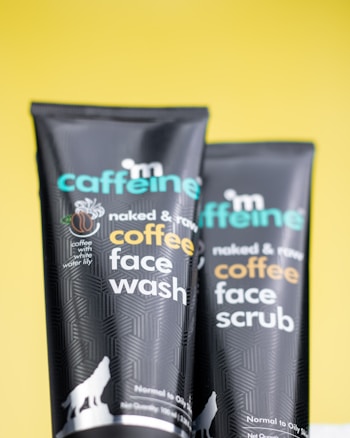 Two black tubes with colorful text display skincare products on a bright yellow background. Each tube features branding details and product descriptions like 'coffee face wash' and 'coffee face scrub'. A small graphic of a wolf at the bottom adds a distinct design element.