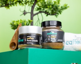 A vibrant setup features skincare products labeled as coffee face mask and coffee body scrub in jars. The jars sit on a green surface with wooden measurement spoons nearby. Behind them, a partially visible small towel with branding is tucked in. A lush green plant provides a natural backdrop.