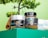 A vibrant setup features skincare products labeled as coffee face mask and coffee body scrub in jars. The jars sit on a green surface with wooden measurement spoons nearby. Behind them, a partially visible small towel with branding is tucked in. A lush green plant provides a natural backdrop.