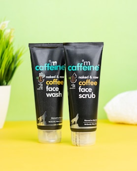 Two black tubes of skincare products are placed on a bright yellow surface against a pale green background. The products are labeled as coffee face wash and coffee face scrub by mCaffeine. A potted plant and a white towel are slightly visible in the background, adding an element of freshness.