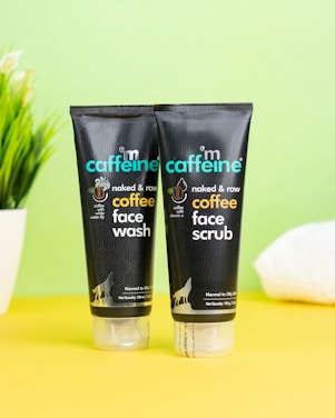 Two black tubes of skincare products are placed on a bright yellow surface against a pale green background. The products are labeled as coffee face wash and coffee face scrub by mCaffeine. A potted plant and a white towel are slightly visible in the background, adding an element of freshness.