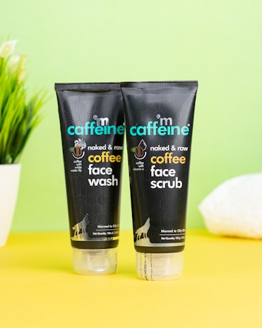 Two black tubes of skincare products are placed on a bright yellow surface against a pale green background. The products are labeled as coffee face wash and coffee face scrub by mCaffeine. A potted plant and a white towel are slightly visible in the background, adding an element of freshness.