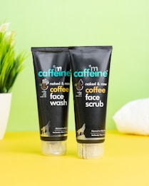 Two black tubes of skincare products are placed on a bright yellow surface against a pale green background. The products are labeled as coffee face wash and coffee face scrub by mCaffeine. A potted plant and a white towel are slightly visible in the background, adding an element of freshness.