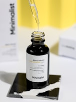 A small black bottle labeled 'Alpha Arbutin 2% Face Serum' sits on a two-tone black and white surface. A dropper is held above the bottle, dispensing a clear liquid. The background features soft yellow and a slightly blurred box with 'Minimalist' written on it.