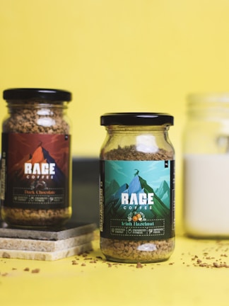 Two glass jars of coffee labeled 'Rage Coffee' with distinct designs and colors are placed against a bright yellow background. The jar on the left is labeled 'Dark Chocolate' and has a dark-themed label, while the jar on the right is labeled 'Irish Hazelnut' with a green and blue mountain design. Coffee granules are scattered around the jars, and a few tiles are placed beneath the left jar.