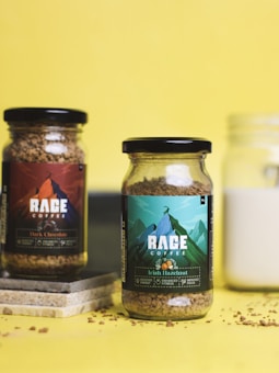Two glass jars of coffee labeled 'Rage Coffee' with distinct designs and colors are placed against a bright yellow background. The jar on the left is labeled 'Dark Chocolate' and has a dark-themed label, while the jar on the right is labeled 'Irish Hazelnut' with a green and blue mountain design. Coffee granules are scattered around the jars, and a few tiles are placed beneath the left jar.