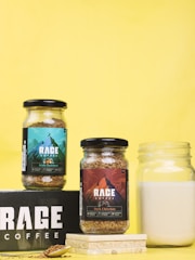 Two jars of Rage Coffee are displayed against a vibrant yellow background. One jar contains Irish Hazelnut coffee, while the other contains Dark Chocolate coffee. The labels feature colorful designs with mountain imagery. A glass jar of milk is placed nearby, along with a spoon and coffee granules on a black Rage Coffee box.