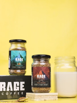 Two jars of Rage Coffee are displayed against a vibrant yellow background. One jar contains Irish Hazelnut coffee, while the other contains Dark Chocolate coffee. The labels feature colorful designs with mountain imagery. A glass jar of milk is placed nearby, along with a spoon and coffee granules on a black Rage Coffee box.