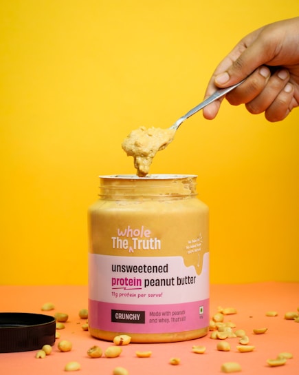 A jar of unsweetened protein peanut butter labeled 'whole The Truth' is partially open with a spoon held above it, scooping out some of the peanut butter. The background is a vibrant yellow, and there are scattered peanuts around the jar.