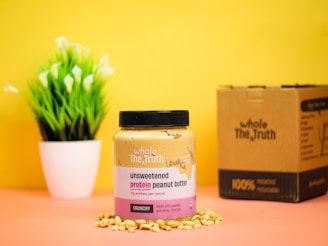 A jar of unsweetened protein peanut butter is placed on a surface surrounded by peanuts, against a bright yellow background. Next to it is a cardboard box, and a small potted plant with green leaves and white flowers is in the background.