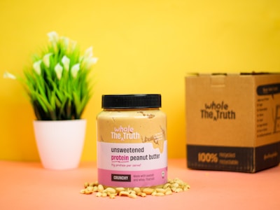 A jar of unsweetened protein peanut butter is placed on a surface surrounded by peanuts, against a bright yellow background. Next to it is a cardboard box, and a small potted plant with green leaves and white flowers is in the background.