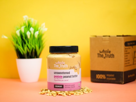 A jar of unsweetened protein peanut butter is placed on a surface surrounded by peanuts, against a bright yellow background. Next to it is a cardboard box, and a small potted plant with green leaves and white flowers is in the background.