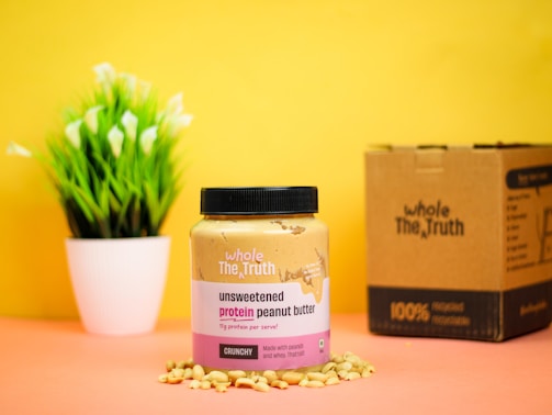 A jar of unsweetened protein peanut butter is placed on a surface surrounded by peanuts, against a bright yellow background. Next to it is a cardboard box, and a small potted plant with green leaves and white flowers is in the background.