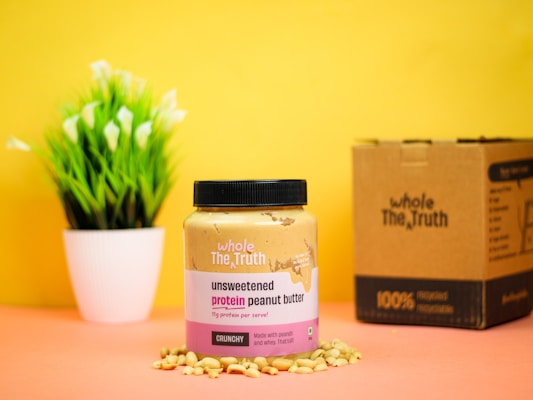 A jar of unsweetened protein peanut butter is placed on a surface surrounded by peanuts, against a bright yellow background. Next to it is a cardboard box, and a small potted plant with green leaves and white flowers is in the background.