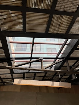 A ceiling with a partially transparent section featuring metal beams and corrugated metal sheets. Sunlight streams through the transparent panels providing a contrast between light and shadow. A wooden beam or light fixture hangs diagonally across the foreground.