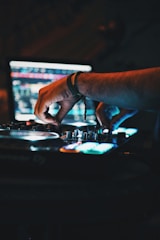 Hands adjusting controls on a DJ mixer with a laptop screen in the background displaying music software interface. The scene is dimly lit, creating a moody atmosphere with colorful lights reflecting off the equipment.