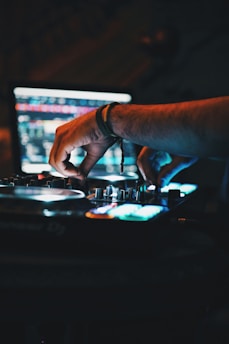 Hands adjusting controls on a DJ mixer with a laptop screen in the background displaying music software interface. The scene is dimly lit, creating a moody atmosphere with colorful lights reflecting off the equipment.