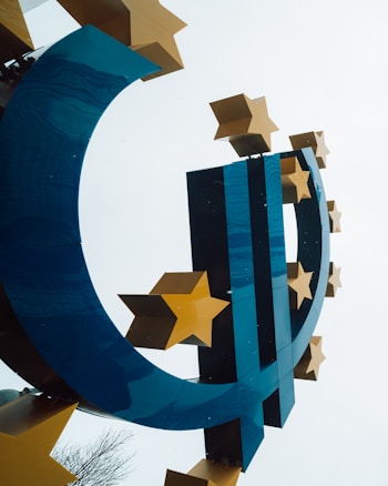 A stylized blue and gold sculpture of the euro currency symbol, adorned with several golden stars. The sculpture stands out against a pale sky background, with a hint of tree branches visible at the base.