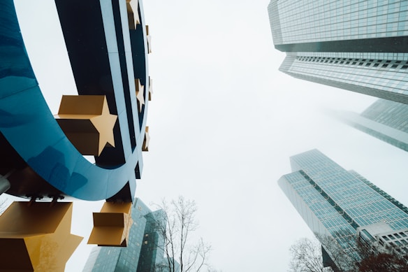 A large blue euro symbol with yellow stars is prominently displayed against the backdrop of modern skyscrapers and a cloudy sky. The buildings are tall and glass-covered, reaching into the mist, creating a sense of height and modern architecture. Bare tree branches are visible at the bottom, adding contrast to the urban scene.