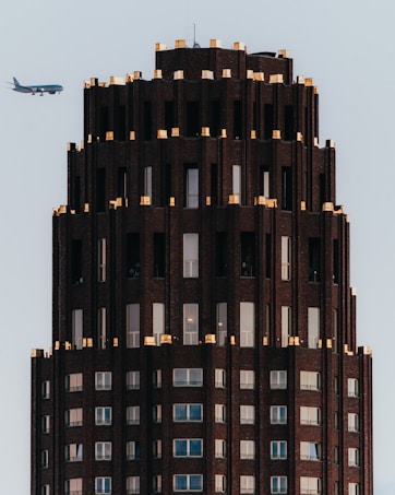 A tall, dark, intricately designed skyscraper with golden accents on the top edges, featuring multiple windows. An airplane is flying in the background against a clear sky.