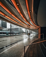 A futuristic urban scene featuring a tram station beneath a modern architectural canopy with illuminated orange lights. The reflection of buildings is visible on the wet pavement after rain, adding a dramatic effect to the cityscape. High-rise office buildings are in the background, contributing to the metropolitan atmosphere.