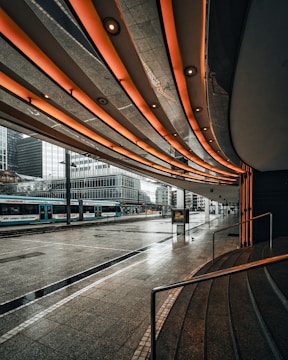 A futuristic urban scene featuring a tram station beneath a modern architectural canopy with illuminated orange lights. The reflection of buildings is visible on the wet pavement after rain, adding a dramatic effect to the cityscape. High-rise office buildings are in the background, contributing to the metropolitan atmosphere.
