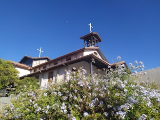 A church building with multiple steeples and crosses on top, surrounded by lush greenery and blooming flowers. The sky is clear and bright blue with a small visible moon during the day.