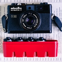 A vintage black Minolta HI-MATIC F camera is positioned above a red container holding several film canisters. The background consists of pages with text, giving the image a nostalgic and vintage feel.