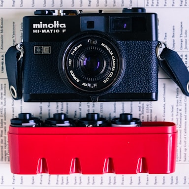 A vintage black Minolta HI-MATIC F camera is positioned above a red container holding several film canisters. The background consists of pages with text, giving the image a nostalgic and vintage feel.