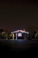 A small, illuminated kiosk stands in a dark area with a person visible inside. The building is adorned with lights along the edges, creating a bright contrast against the night. Surrounding the kiosk are stacked chairs and a few plants in pots. Buildings and a crane are faintly visible in the background.