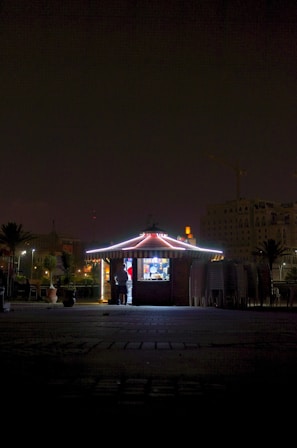 A small, illuminated kiosk stands in a dark area with a person visible inside. The building is adorned with lights along the edges, creating a bright contrast against the night. Surrounding the kiosk are stacked chairs and a few plants in pots. Buildings and a crane are faintly visible in the background.