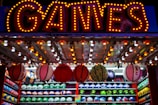 A brightly lit carnival game stall with a large 'GAMES' sign at the top surrounded by red and yellow lights. Various sports balls, including basketballs and soccer balls, are hanging from the ceiling and neatly arranged on shelves below.