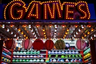 A brightly lit carnival game stall with a large 'GAMES' sign at the top surrounded by red and yellow lights. Various sports balls, including basketballs and soccer balls, are hanging from the ceiling and neatly arranged on shelves below.