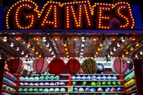A brightly lit carnival game stall with a large 'GAMES' sign at the top surrounded by red and yellow lights. Various sports balls, including basketballs and soccer balls, are hanging from the ceiling and neatly arranged on shelves below.