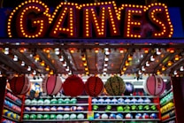 A brightly lit carnival game stall with a large 'GAMES' sign at the top surrounded by red and yellow lights. Various sports balls, including basketballs and soccer balls, are hanging from the ceiling and neatly arranged on shelves below.