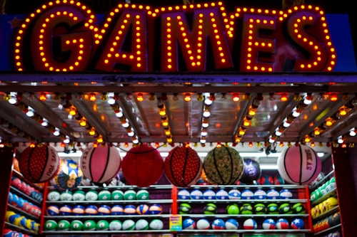 A brightly lit carnival game stall with a large 'GAMES' sign at the top surrounded by red and yellow lights. Various sports balls, including basketballs and soccer balls, are hanging from the ceiling and neatly arranged on shelves below.