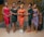 Five women stand together indoors, each wearing vibrant, elegant dresses of different colors. They display a joyful and confident demeanor, with a decorated wall in the background and a large plant to the side.