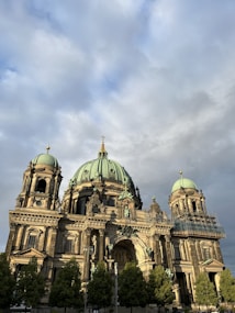 A grand historical building with large, ornate green domes and intricate architectural details stands against a backdrop of a cloudy sky. The structure features columns and statues, with scaffolding visible on one part of the building. Green trees line the front of the building, adding an element of nature to the scene.