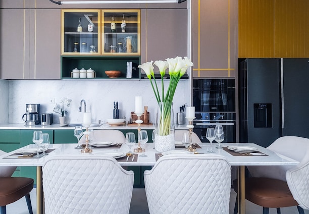 A modern kitchen and dining area featuring a marble-topped table set for four with plates, glasses, and candlesticks. A vase of white flowers is placed at the center. Behind the table, a countertop holds various kitchen appliances like a coffee maker and utensils. Cabinets in muted colors and a large refrigerator are visible.