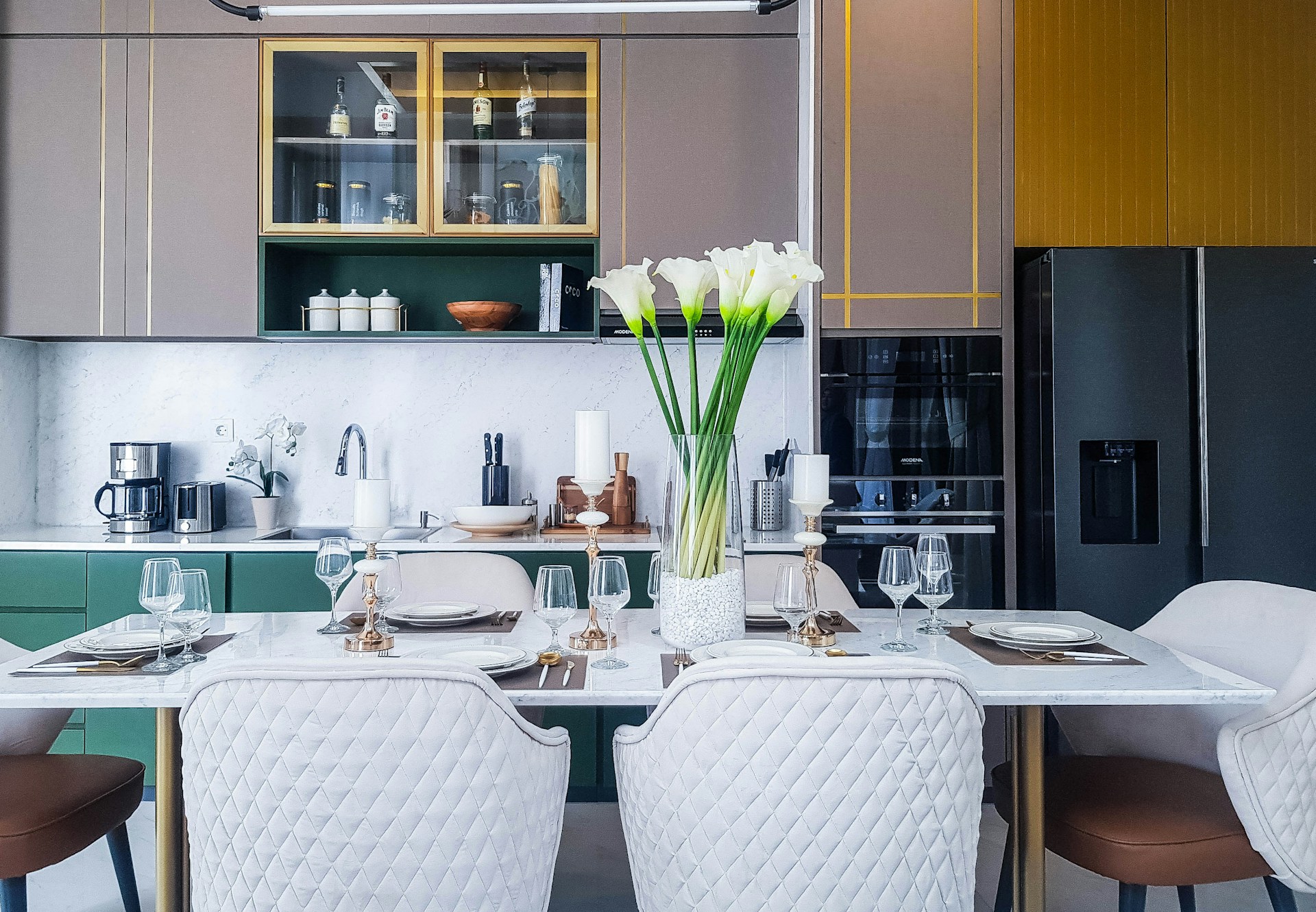 A modern kitchen and dining area featuring a marble-topped table set for four with plates, glasses, and candlesticks. A vase of white flowers is placed at the center. Behind the table, a countertop holds various kitchen appliances like a coffee maker and utensils. Cabinets in muted colors and a large refrigerator are visible.