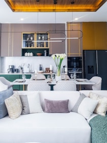 A contemporary kitchen and living area featuring a modern dining table set with wine glasses, plates, and cutlery. The kitchen is equipped with sleek cabinetry, a refrigerator, and decorative items like bottles and bowls. Above the dining area, stylish lighting fixtures can be seen. A white sofa with numerous decorative cushions in various textures is in the foreground, while tall white flowers add an elegant touch.