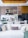 A contemporary kitchen and living area featuring a modern dining table set with wine glasses, plates, and cutlery. The kitchen is equipped with sleek cabinetry, a refrigerator, and decorative items like bottles and bowls. Above the dining area, stylish lighting fixtures can be seen. A white sofa with numerous decorative cushions in various textures is in the foreground, while tall white flowers add an elegant touch.