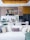 A contemporary kitchen and living area featuring a modern dining table set with wine glasses, plates, and cutlery. The kitchen is equipped with sleek cabinetry, a refrigerator, and decorative items like bottles and bowls. Above the dining area, stylish lighting fixtures can be seen. A white sofa with numerous decorative cushions in various textures is in the foreground, while tall white flowers add an elegant touch.
